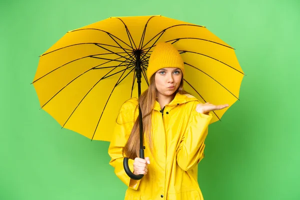 年轻的金发女郎 身穿防雨外套 带着雨伞 盖在孤立的彩色关键背景上 举手表决时疑虑重重 — 图库照片