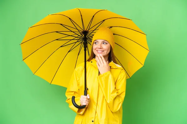 若いですブロンド女性とともにレインプルーフコートと傘上の隔離されたクロマキー背景上の見上げます上の笑顔 — ストック写真