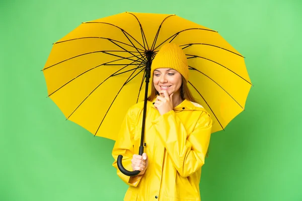 若いですブロンド女性とともにレインプルーフコートと傘上の隔離されたクロマキー背景見ますザ側と笑顔 — ストック写真