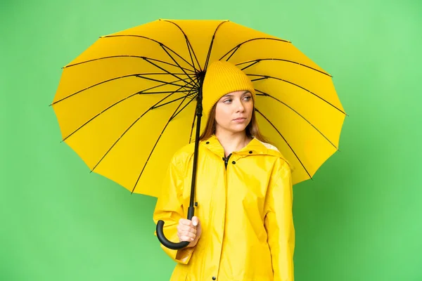 若いですブロンド女性とともにレインプルーフコートと傘上の隔離されたクロマキー背景見ますザ側 — ストック写真