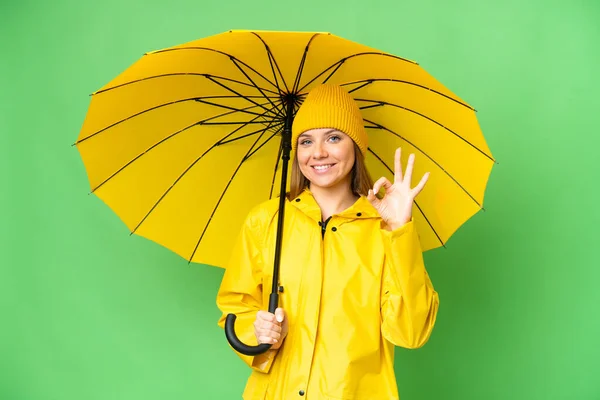 若いですブロンド女性とともにレインプルーフコートと傘上の隔離されたクロマキー背景ショーOk Signとともに指 — ストック写真