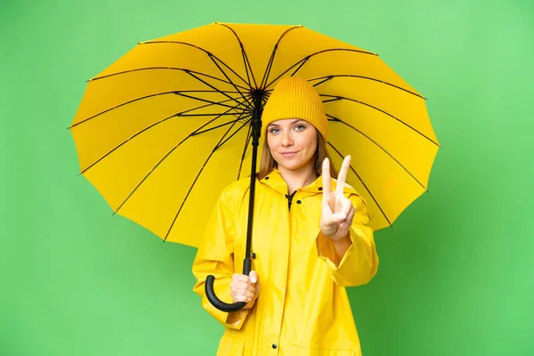 若いですブロンド女性とともにレインプルーフコートと傘上の隔離されたクロマキー背景笑顔とショー勝利サイン — ストック写真
