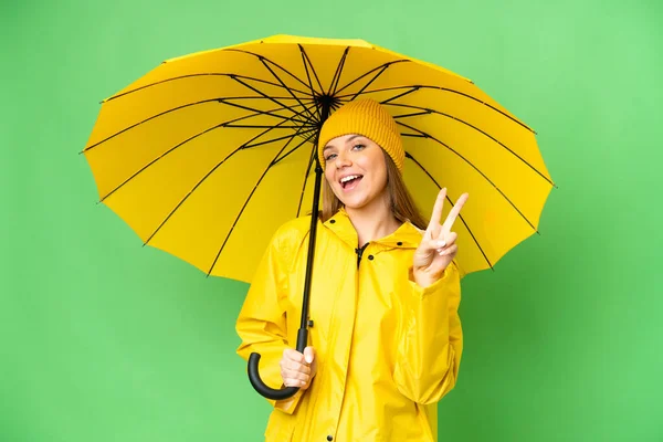 若いですブロンド女性とともにレインプルーフコートと傘上の隔離されたクロマキー背景笑顔とショー勝利サイン — ストック写真