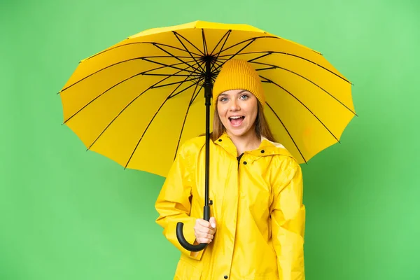 若いですブロンド女性とともにレインプルーフコートと傘上の隔離されたクロマキー背景とともに驚きの表情 — ストック写真