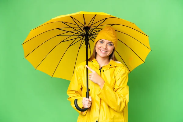若いですブロンド女性とともにレインプルーフコートと傘上の隔離されたクロマキー背景を指す側に製品を提示 — ストック写真