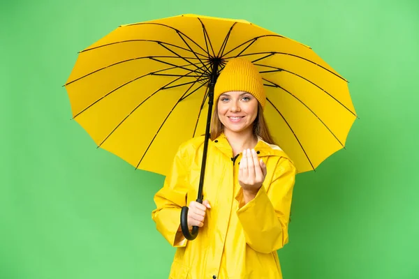 絶縁されたクロマキーの背景にレインプルーフコートと傘を着た若いブロンドの女性が手で来るように誘う 来てくれて嬉しい — ストック写真