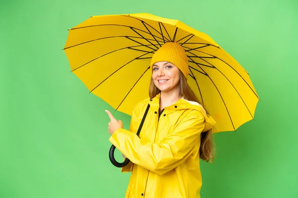 若いですブロンド女性とともにレインプルーフコートと傘上の隔離されたクロマキー背景ポインティングバック — ストック写真