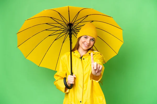 若いですブロンド女性とともにレインプルーフコートと傘上の隔離されたクロマキー背景ショーと持ち上げる指 — ストック写真