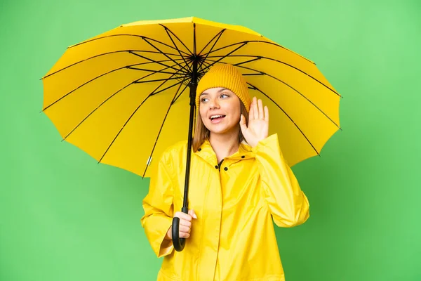 若いですブロンド女性とともにレインプルーフコートと傘上の隔離されたクロマキー背景リスニング何かに手を置くことによって耳 — ストック写真
