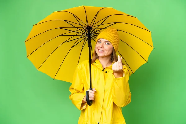 若いですブロンド女性とともにレインプルーフコートと傘上の隔離されたクロマキー背景作りお金ジェスチャー — ストック写真