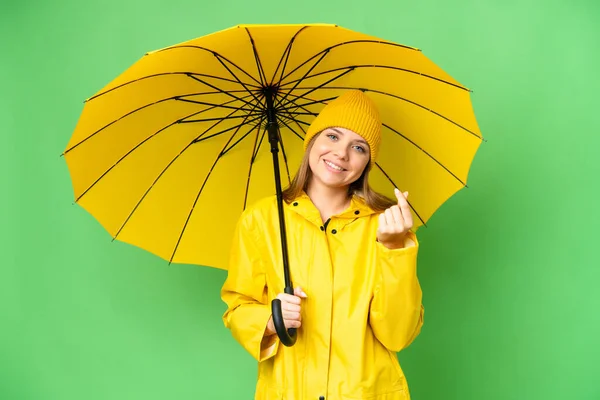 若いですブロンド女性とともにレインプルーフコートと傘上の隔離されたクロマキー背景作りお金ジェスチャー — ストック写真