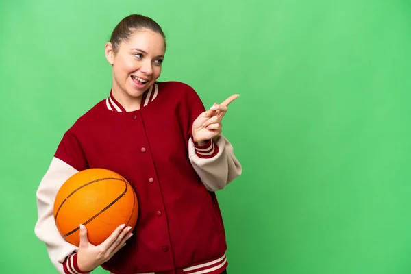 年轻的金发女子在一个孤立的彩色关键背景下打篮球 手指指向旁边 展示自己的产品 — 图库照片