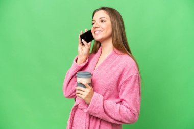 İzole edilmiş krom arka planda bornozlu genç bir kadın. Elinde kahve ve cep telefonu var.