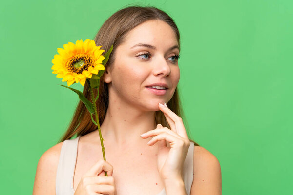 Young blonde woman over isolated chroma key background holding a sunflower while smiling. Close up portrait
