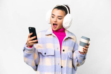 İzole edilmiş beyaz arka planda kışlık kuklalar takan genç bir kadın. Elinde kahve ve cep telefonu var.