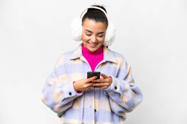 İzole edilmiş beyaz arka plan üzerine kış maskesi takan genç bir kadın cep telefonuyla mesaj gönderiyor.