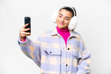İzole edilmiş beyaz arka planda kış örtüsü takan genç bir kadın selfie çekiyor.