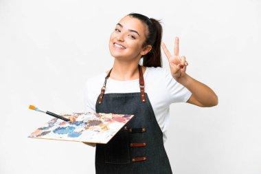 İzole edilmiş beyaz arka planda palet tutan genç sanatçı gülümsüyor ve zafer işareti gösteriyor.