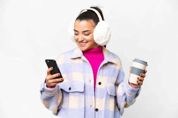Young woman wearing winter muffs over isolated white background holding coffee to take away and a mobile