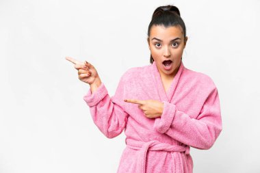 İzole edilmiş beyaz arka planı olan bornozlu genç bir kadın şaşırmış ve işaret ediyor.