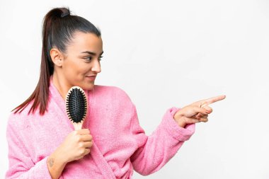 Bornozlu genç bir kadın, saçı taranmış, izole edilmiş beyaz arka planda bir ürün sunmak için kenarı işaret ediyor.