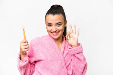 Bornozlu genç bir kadın izole edilmiş beyaz arka planda dişlerini fırçalıyor. Parmaklarıyla işaret veriyor.