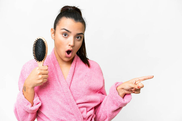 Young woman in a bathrobe with hair comb over isolated white background surprised and pointing side