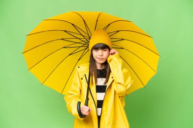 Yağmura dayanıklı ceketi ve şemsiyesi olan küçük bir kız izole edilmiş krom anahtar arka planı hayal kırıklığına uğramış ve kulaklarını kapatmış.