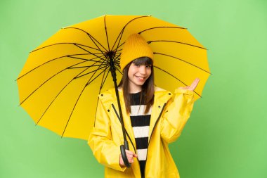 Yağmura dayanıklı ceketi ve şemsiyesi olan küçük bir kız izole edilmiş krom anahtar arka planda. Davet etmek için ellerini yana uzatıyor.