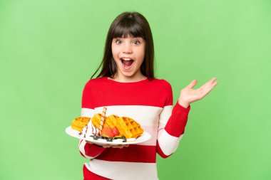 İzole edilmiş krom arka plan üzerinde waffle tutan küçük kız şok olmuş yüz ifadesi ile