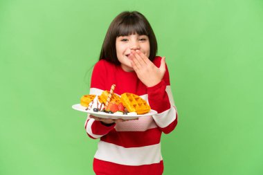 İzole edilmiş krom arka planda waffle tutan küçük bir kız. Mutlulukla gülümsüyor ve ağzı elle örtüyor.