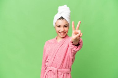 Üzerinde bornoz olan küçük bir kız izole edilmiş krom anahtar arka planda gülümsüyor ve zafer işareti gösteriyor.