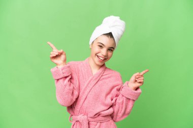 İzole edilmiş krom arka planda bornoz giymiş küçük bir kız parmak uçlarını işaret ediyor ve mutlu.