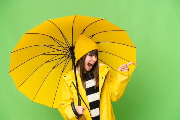 小さな女の子とともにレインプルーフコートと傘上の隔離されたクロマキー背景指差し指で側面と製品を提示 — ストック写真
