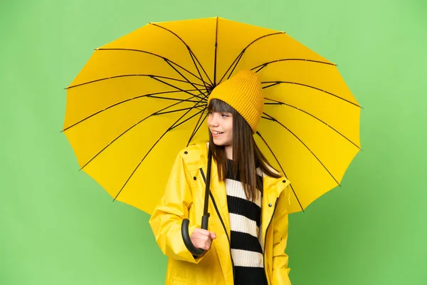 小さな女の子とともにレインプルーフコートと傘上の隔離されたクロマキー背景見ますザ側と笑顔 — ストック写真