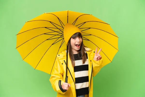 小さな女の子とともにレインプルーフコートと傘上の隔離されたクロマキー背景笑顔とショー勝利サイン — ストック写真