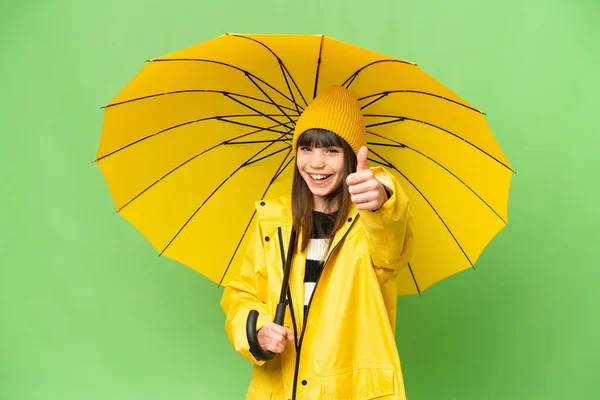 小さな女の子ですレインプルーフコートと傘上の隔離されたクロマキー背景上の親指アップとともに何か良いことが起こっているので — ストック写真