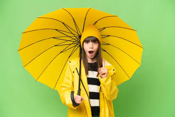 指を持ち上げながら解決策を実現しようとしている隔離されたクロマキーの背景にレインプルーフコートと傘を持つ小さな女の子 — ストック写真