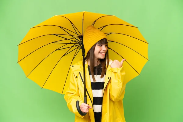 レインプルーフコートと傘を持つ小さな女の子上の隔離されたクロマキー背景を指す側に製品を提示 — ストック写真