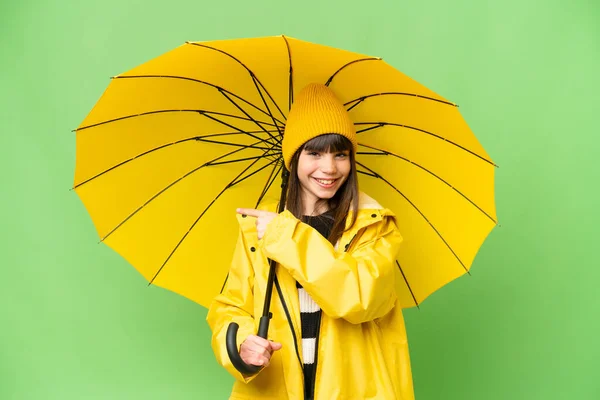 レインプルーフコートと傘を持つ小さな女の子上の隔離されたクロマキー背景を指す側に製品を提示 — ストック写真