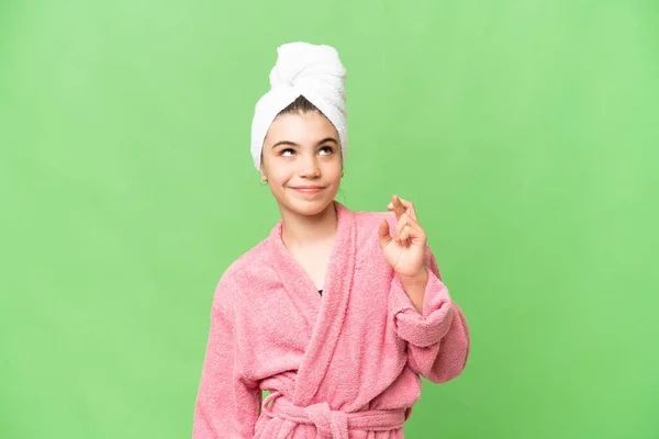 Little girl in a bathrobe over isolated chroma key background with fingers crossing and wishing the best
