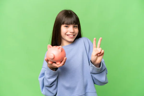 Kleines Mädchen Mit Sparschwein Über Isoliertem Chroma Schlüsselhintergrund Lächelt Und — Stockfoto