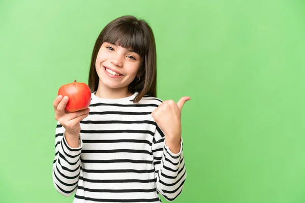 Little girl with an apple over isolated chroma key background pointing to the side to present a product