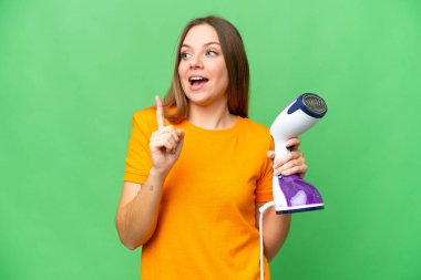 Sarışın genç bir kadın izole edilmiş krom anahtar arkaplan üzerinde dikey buhar ütüsü tutuyor. Çözümü fark etmek için parmağını kaldırıyor.