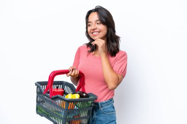 Elinde yiyecek dolu bir alışveriş sepeti olan genç bir kadın yana bakıyor ve gülümsüyor.