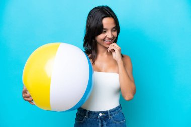 Mavi arka planda izole edilmiş plaj topu tutan genç İspanyol kadın yana bakıyor ve gülümsüyor.