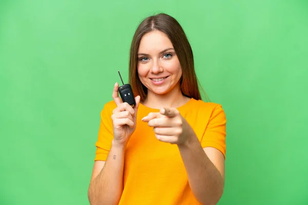Young woman holding car keys over isolated chroma key background points finger at you with a confident expression