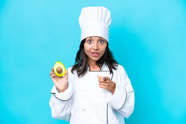 Young hispanic chef woman holding avocado isolated on blue background surprised and pointing front