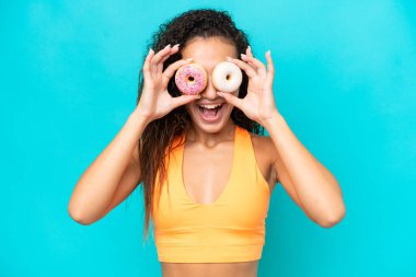 Genç bir Arap kadın mavi arka planda gözünün içine donut tıkıştırıyor.