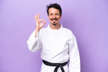 Mor arka planda karate yapan genç beyaz adam parmaklarıyla tamam işareti gösteriyor.